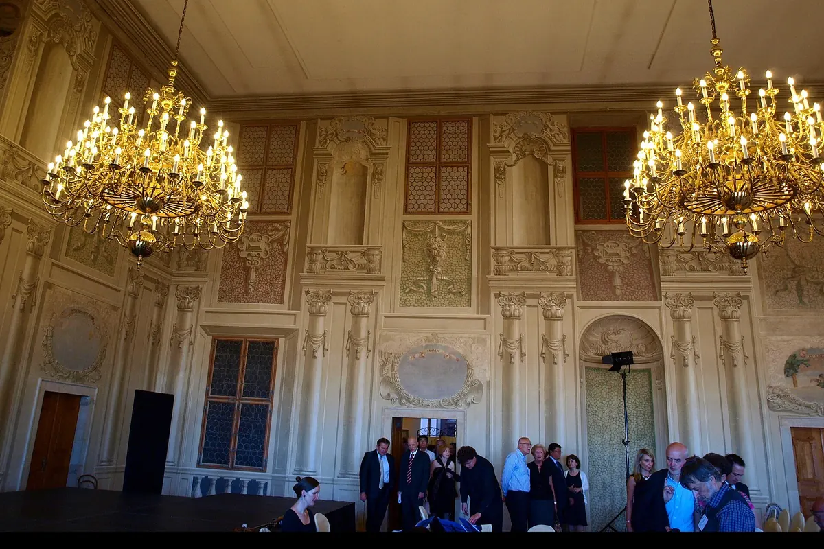 A hall of the Lobkowicz Palace with two gilded chandeliers, painted wall panels and rows of chairs set out for a performance.