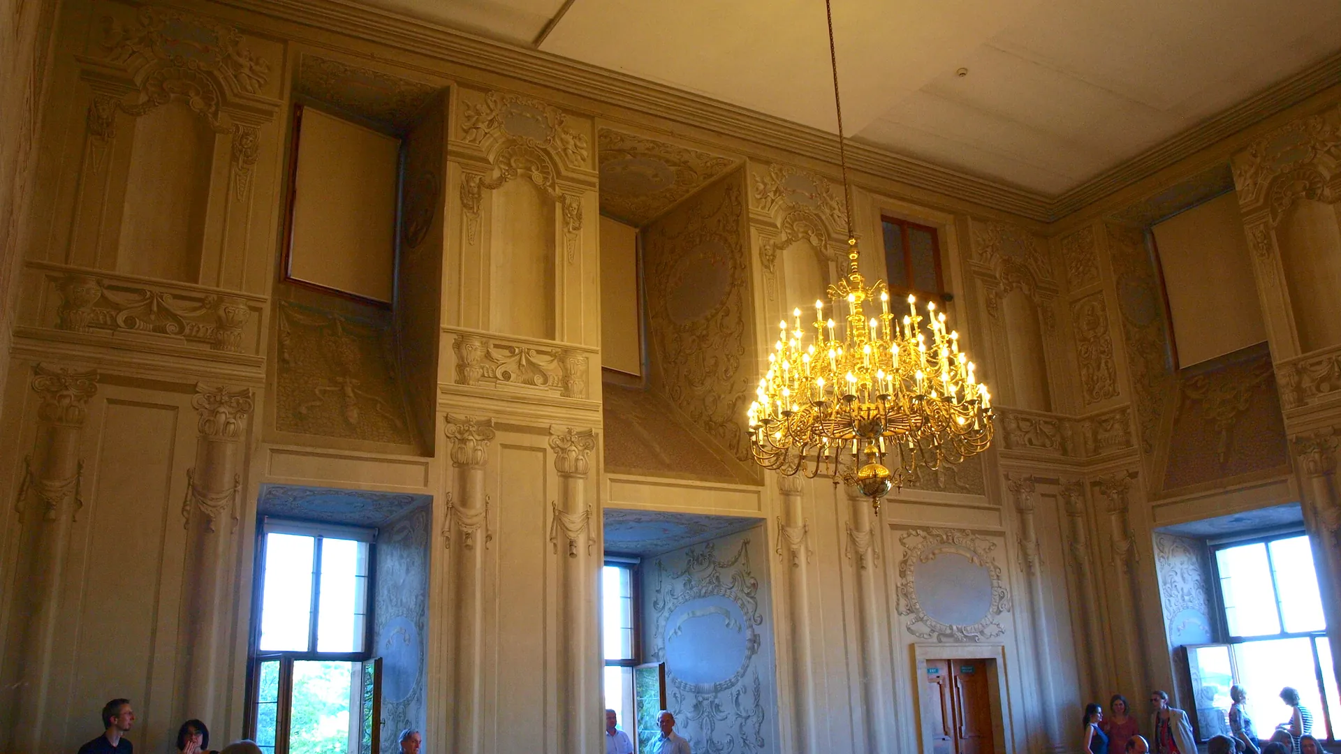 A tall hall in Lobkowicz Palace with painted architectural panels on the walls, a lit gilded chandelier at the right and tall windows below.