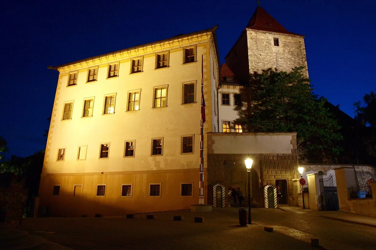 The Lobkowicz Palace lit up at night on the castle hill, with a street lamp and a tower behind it.